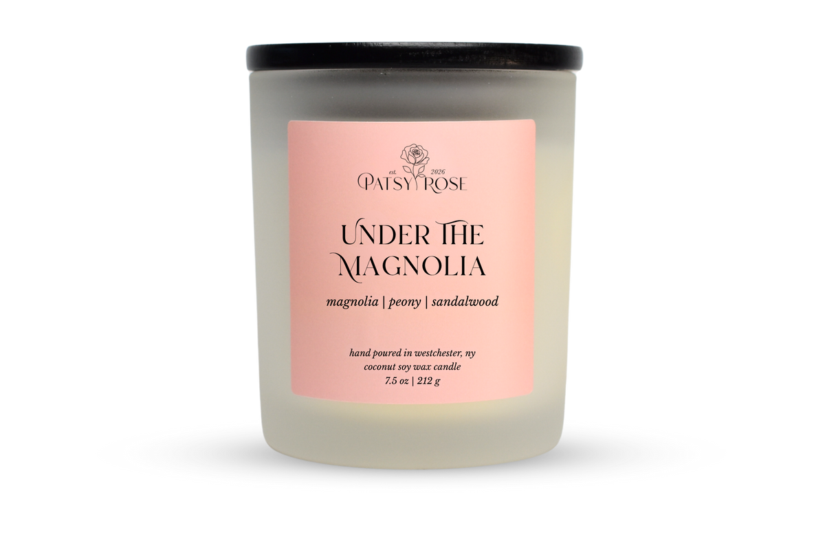 A 7.5 oz soy wax candle by Patsy Rose in the 'Under The Magnolia' 'magnolia | peony | sandalwood' scent. The candle is in a frosted glass jar with a black lid and a peach-pink label, set against a plain white background.