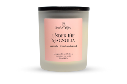 A 7.5 oz soy wax candle by Patsy Rose in the 'Under The Magnolia' 'magnolia | peony | sandalwood' scent. The candle is in a frosted glass jar with a black lid and a peach-pink label, set against a plain white background.