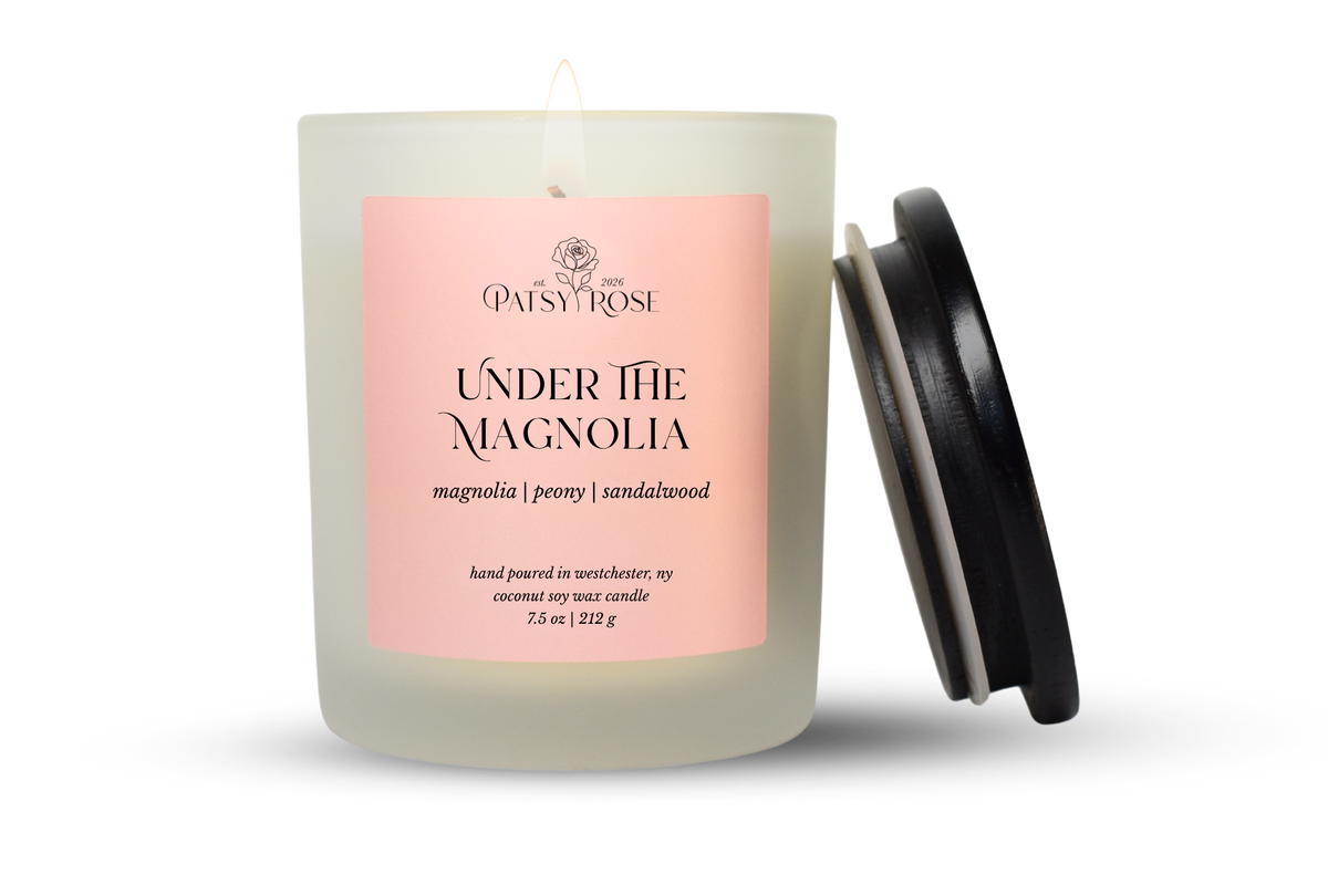 A 7.5 oz single-wick candle in a frosted glass jar with a pale pink label reading 'Patsy Rose Under The Magnolia' and 'magnolia | peony | sandalwood.' A black lid is leaning against the right side of the jar on a clean white background.