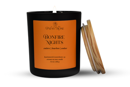 A single-wick coconut soy candle in a matte black glass jar with an orange label reading 'Patsy Rose Bonfire Nights' and 'embers | bourbon | amber.' A wood lid leans against the right side of the jar on a clean white background.