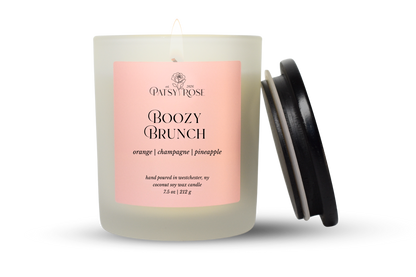 A 7.5 oz single-wick candle in a frosted glass jar with a pale pink label reading 'Patsy Rose Boozy Brunch' and 'orange | champagne | pineapple.' A black lid is leaning against the right side of the jar on a clean white background.
