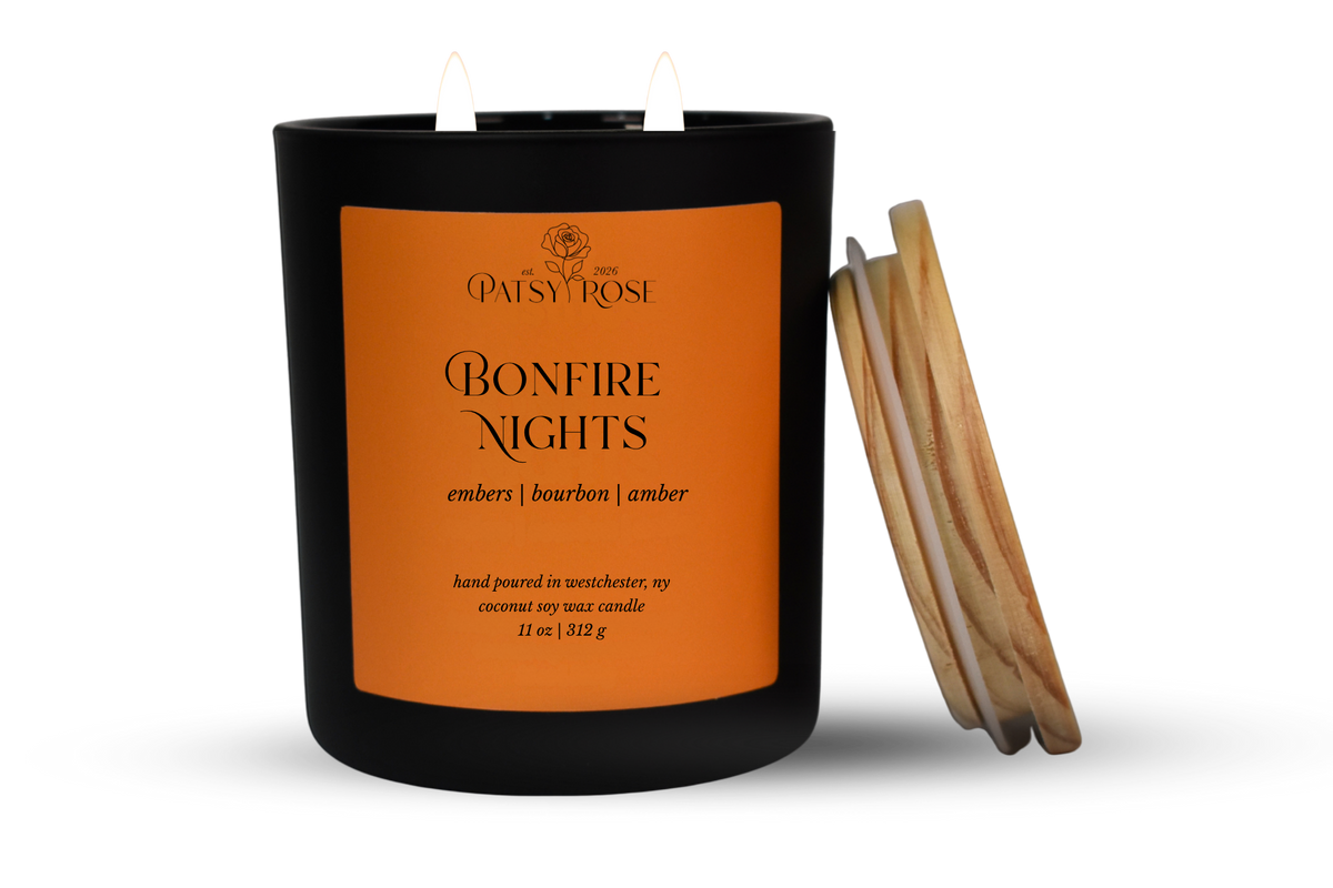 An 11 oz double-wick candle in a matte black glass jar with an orange label reading 'Patsy Rose Bonfire Nights' and 'embers | bourbon | amber.' A wood lid is leaning against the right side of the jar on a clean white background.