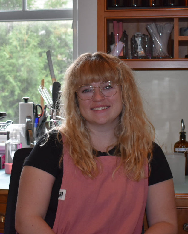 A smiling woman with blonde hair and glasses sits in a candle craft workspace for Patsy Rose with wooden cabinets, shelves of bottles, and candle making equipment in the background.